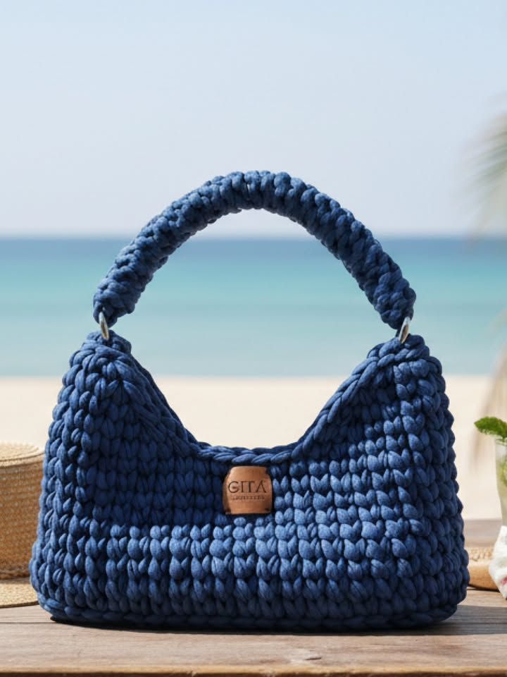 Bolsa de crochê azul-marinho em fio de malha grosso, formato baguete com alça arredondada e etiqueta de couro, sobre madeira à beira-mar