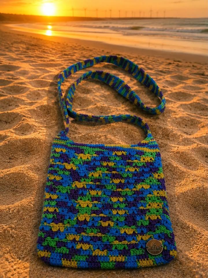 Bolsa transversal de crochê multicolor em azul, verde e amarelo, com alça longa, na areia ao pôr do sol